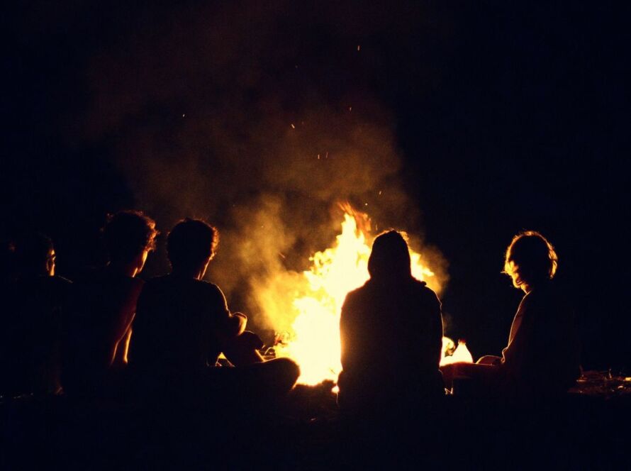 Ilustração de um acampamento pré-histórico à noite, com uma fogueira ao centro, um caçador em silêncio e lobos observando à distância, na penumbra da floresta.